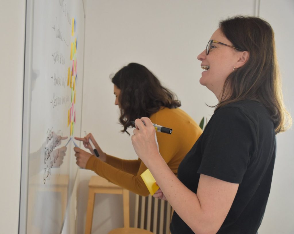 Dos profesionales de EINNA impartiendo una formación en sostenibilidad: escriben sobre una pizarra blanca llena de notas adhesivas de colores, y una de ellas sonríe.